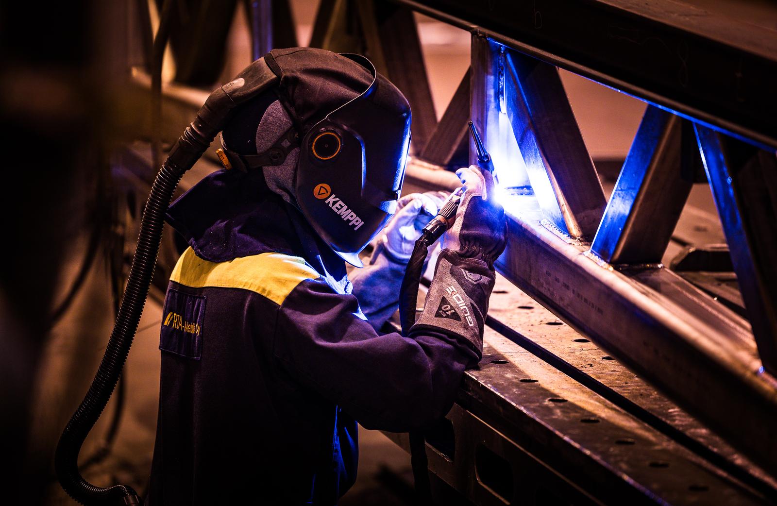 Hitsauksen ammattilainen työskentelemässä metallirakenteen parissa suojavarusteet yllään. Kirkas valokaari valaisee hitsauskohdan.