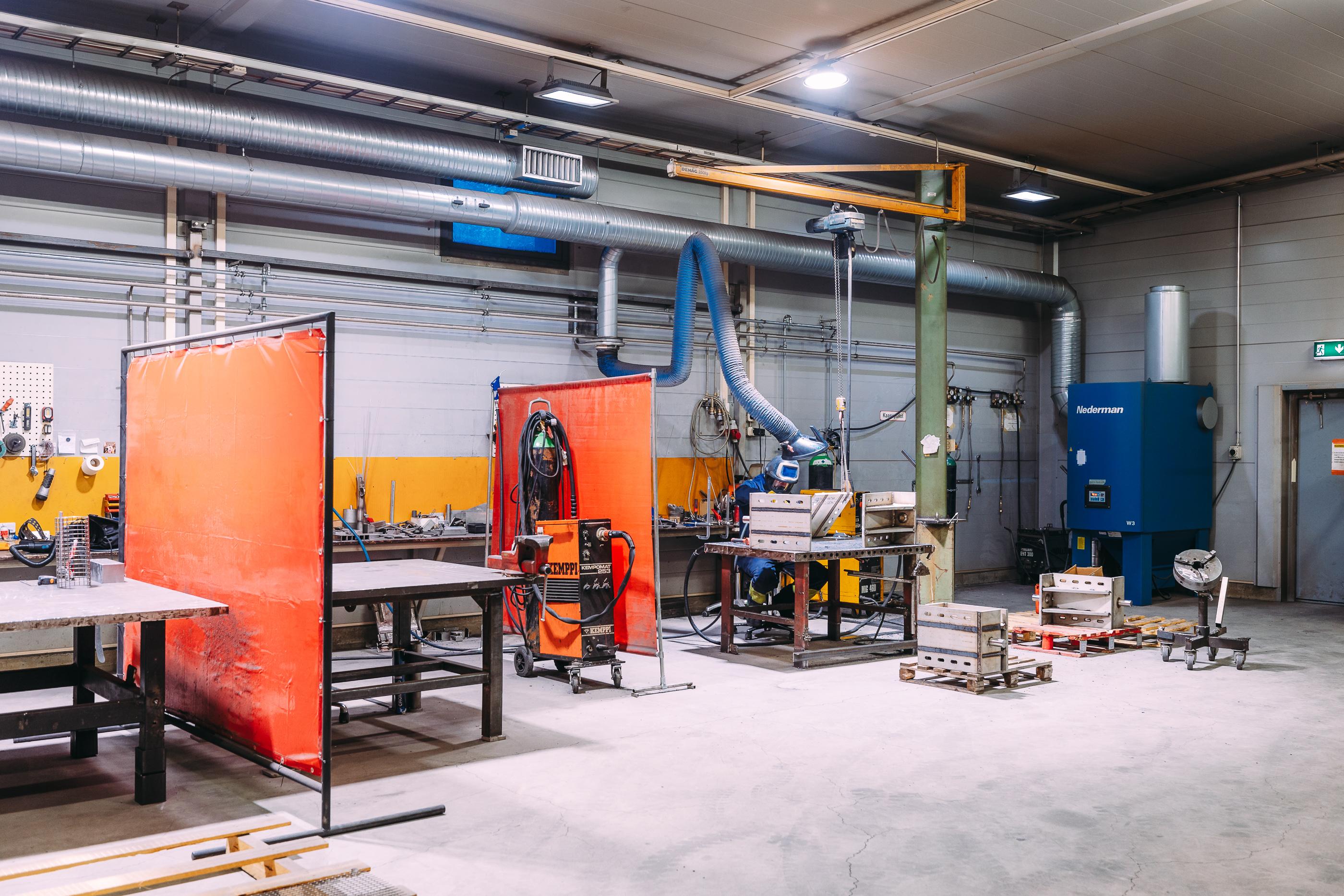 Teollinen kokoonpanotila, jossa on hitsaus- ja kokoonpanopisteit&auml;. Kuvassa n&auml;kyy ty&ouml;p&ouml;yti&auml;, suojaverhoja, erilaisia ty&ouml;kaluja sek&auml; ilmanvaihtoj&auml;rjestelm&auml;, joka varmistaa turvallisen ty&ouml;skentely-ymp&auml;rist&ouml;n.