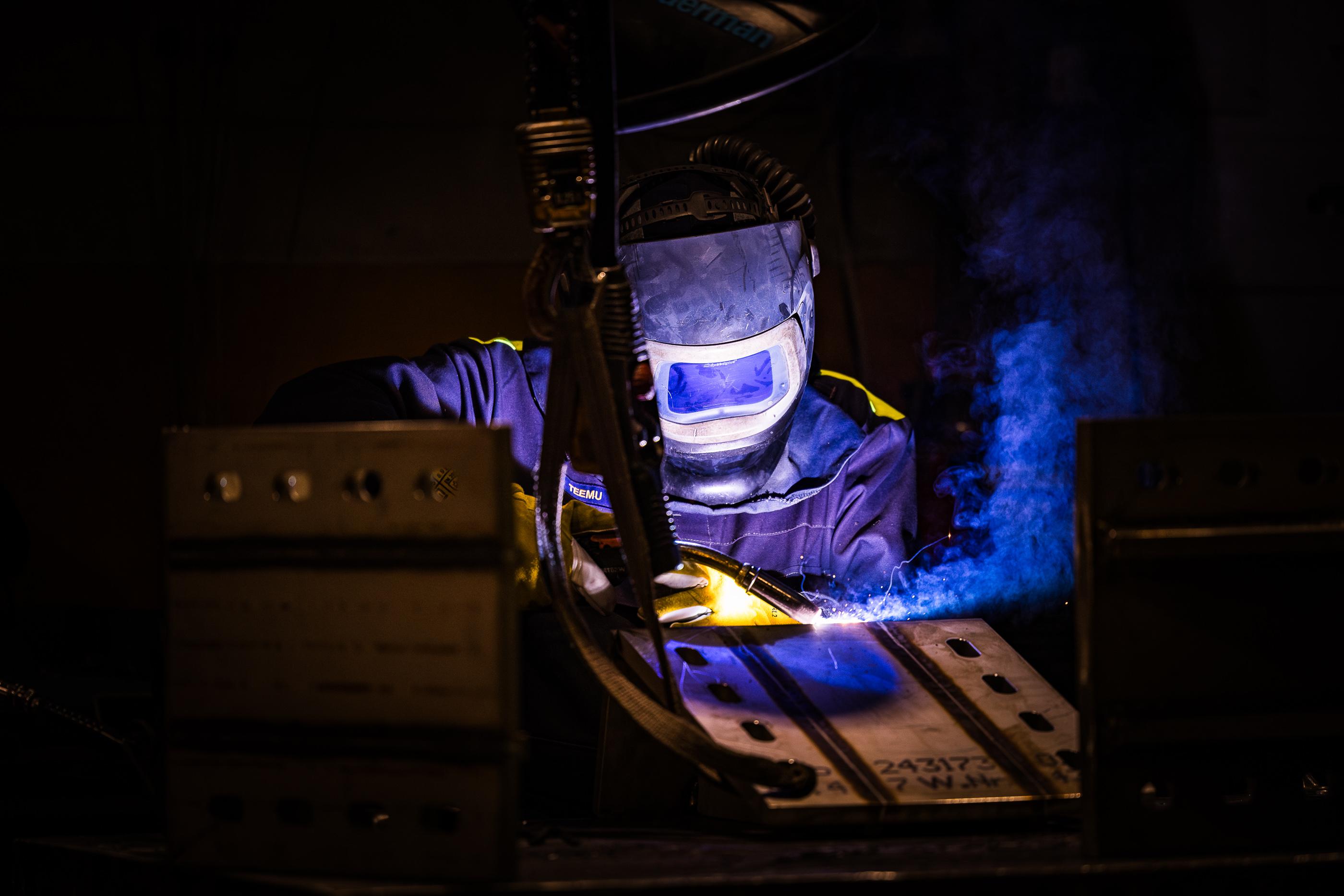 Hitsausammattilainen ty&ouml;skentelee suojavarusteissa, hitsaten metallirakennetta tarkasti. Hitsausprosessissa syntyy sininen valokaari ja savua.