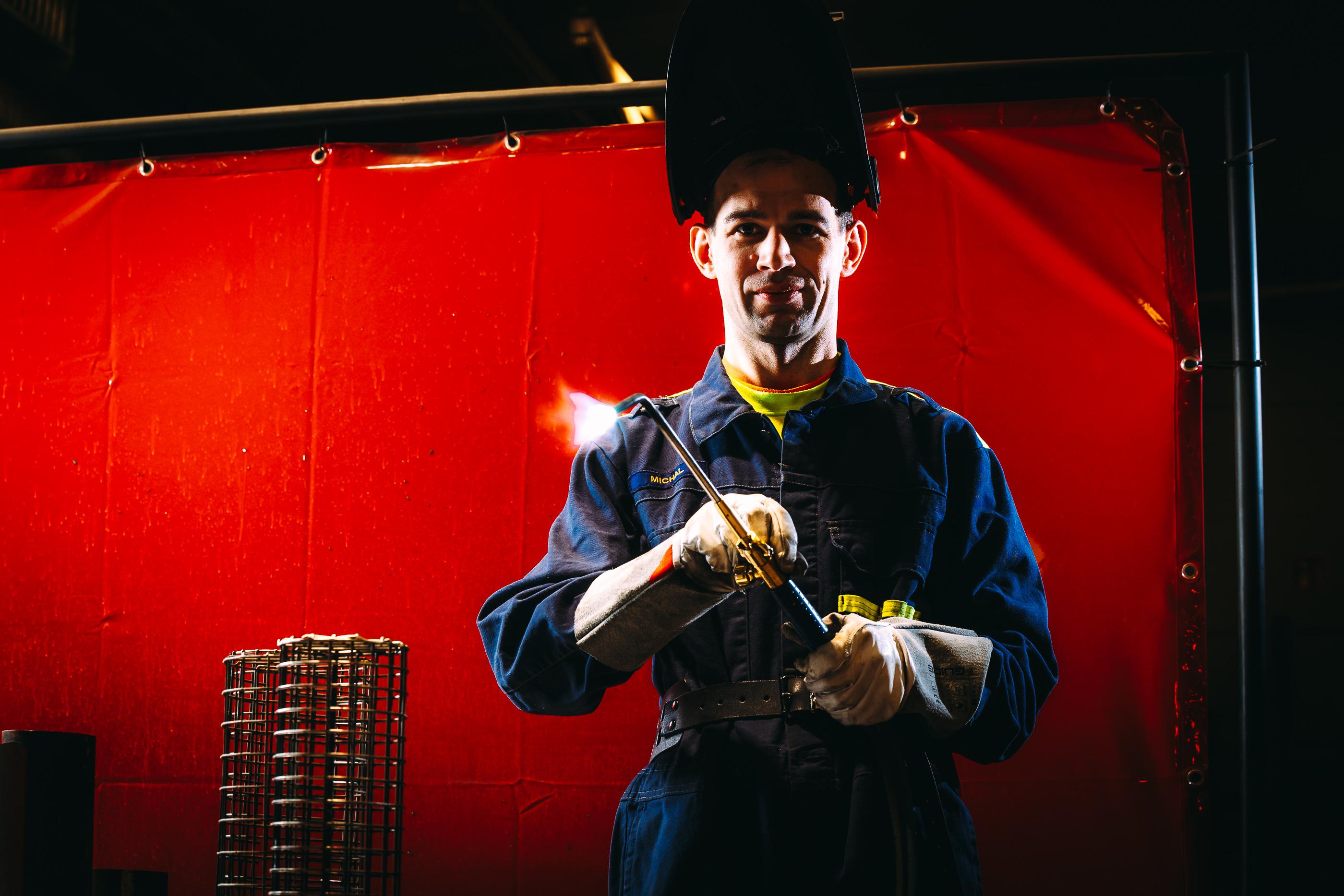 A welding professional wearing work clothes holds a gas torch in front of a red protective curtain.
