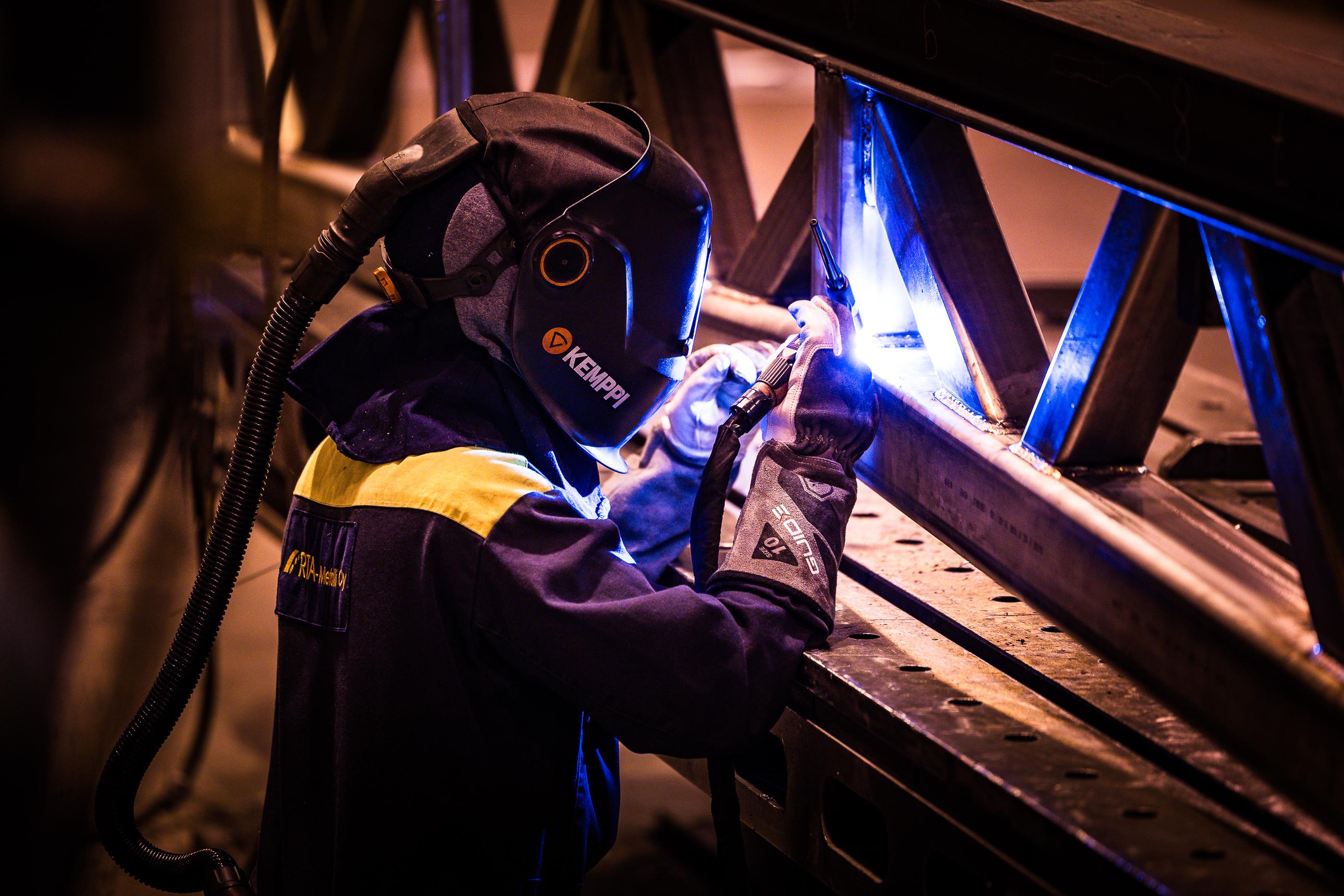 Hitsausammattilainen suorittaa tarkkuushitsausta metallirakenteessa, suojavarusteet p&auml;&auml;ll&auml; ja kirkas hitsausvalo ymp&auml;rill&auml;&auml;n.