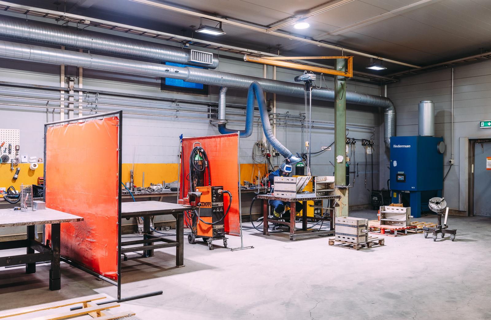 Teollinen kokoonpanotila, jossa on hitsaus- ja kokoonpanopisteitä. Kuvassa näkyy työpöytiä, suojaverhoja, erilaisia työkaluja sekä ilmanvaihtojärjestelmä, joka varmistaa turvallisen työskentely-ympäristön.